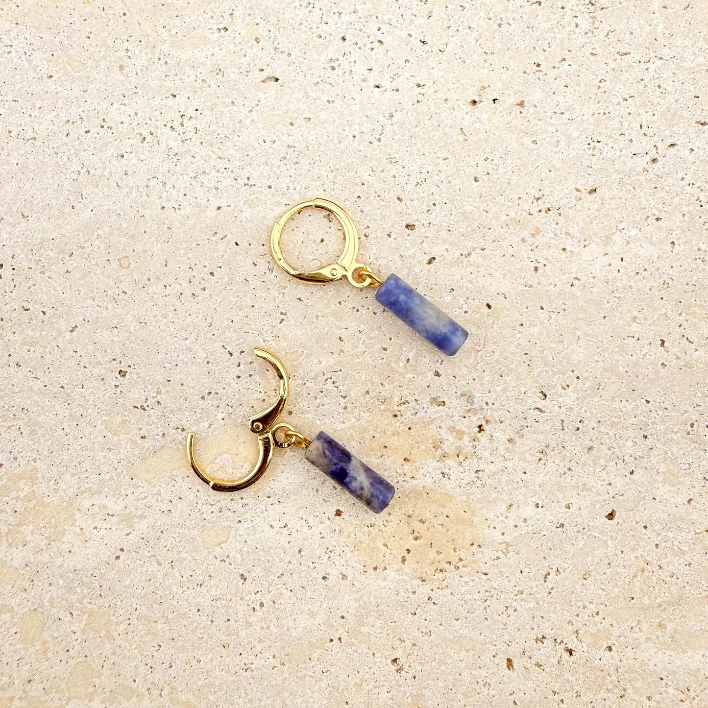 A pair of gold Aking Sinta huggie hoop earrings featuring blue sodalite column charms. One earring is shown with the clasp open to demonstrate the hinge mechanism. The natural blue and white marbled gemstones hang from polished gold hoops on a light natural stone surface