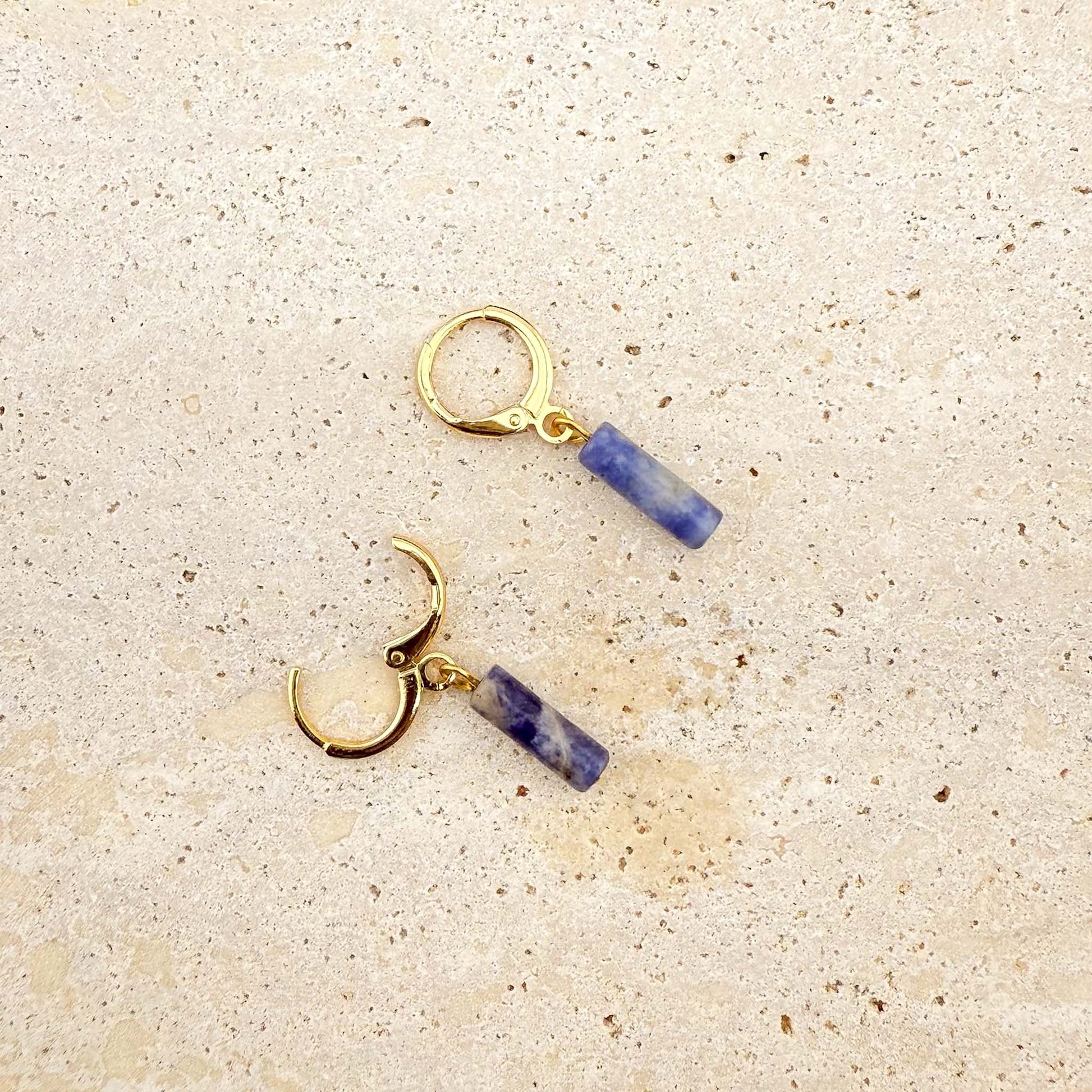 A pair of gold Aking Sinta huggie hoop earrings featuring blue sodalite column charms. One earring is shown with the clasp open to demonstrate the hinge mechanism. The natural blue and white marbled gemstones hang from polished gold hoops on a light natural stone surface