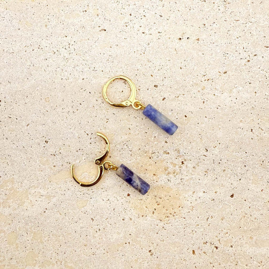 A pair of gold Aking Sinta huggie hoop earrings featuring blue sodalite column charms. One earring is shown with the clasp open to demonstrate the hinge mechanism. The natural blue and white marbled gemstones hang from polished gold hoops on a light natural stone surface