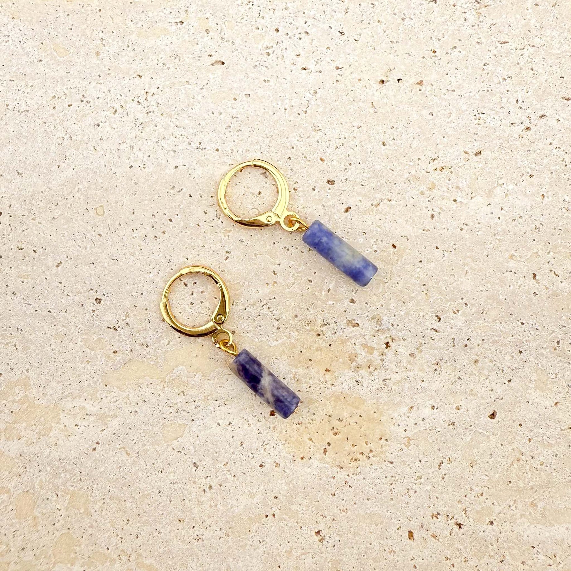 A pair of Aking Sinta gold huggie hoop earrings featuring blue sodalite column charms. The natural gemstone cylinders show unique marbled patterns of deep blue and white, suspended from polished gold-toned hoops on a light natural stone surface.