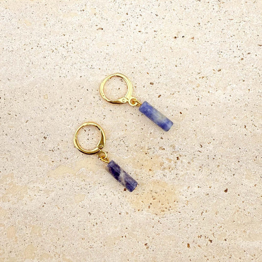 A pair of Aking Sinta gold huggie hoop earrings featuring blue sodalite column charms. The natural gemstone cylinders show unique marbled patterns of deep blue and white, suspended from polished gold-toned hoops on a light natural stone surface.