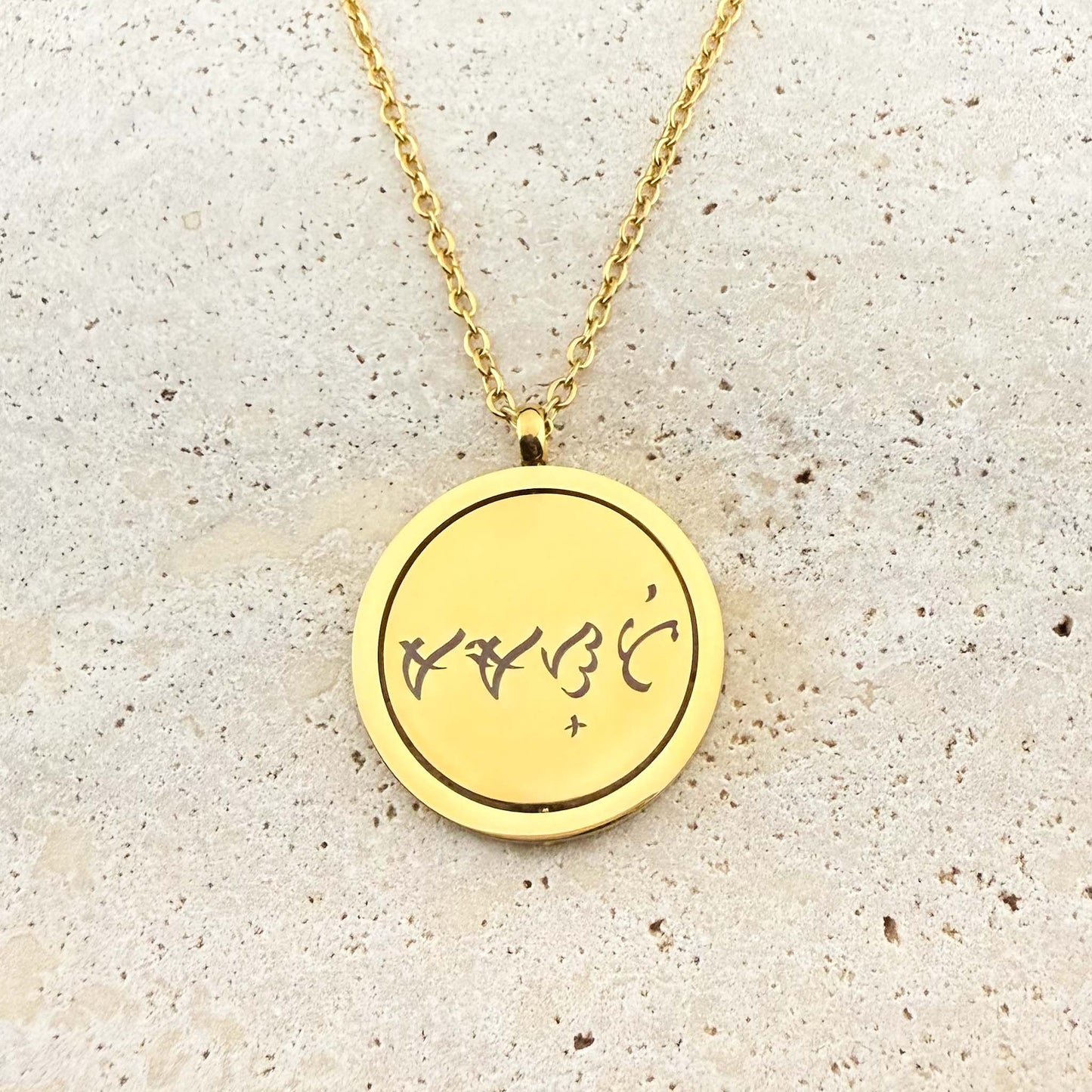 A close-up of an Aking Sinta gold spinning coin fidget necklace. The circular pendant features a bespoke engraving of ancient Filipino Baybayin script on a polished gold surface. The pendant hangs from a delicate gold cable chain and is displayed on a light, textured stone background