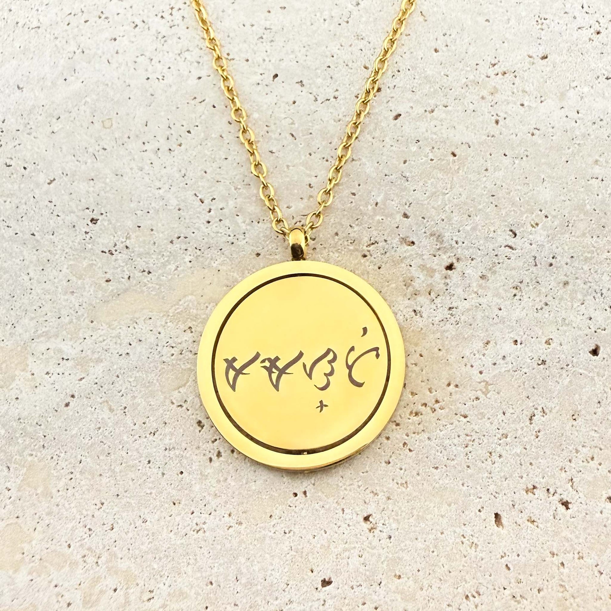 A close-up of an Aking Sinta gold spinning coin fidget necklace. The circular pendant features a bespoke engraving of ancient Filipino Baybayin script on a polished gold surface. The pendant hangs from a delicate gold cable chain and is displayed on a light, textured stone background