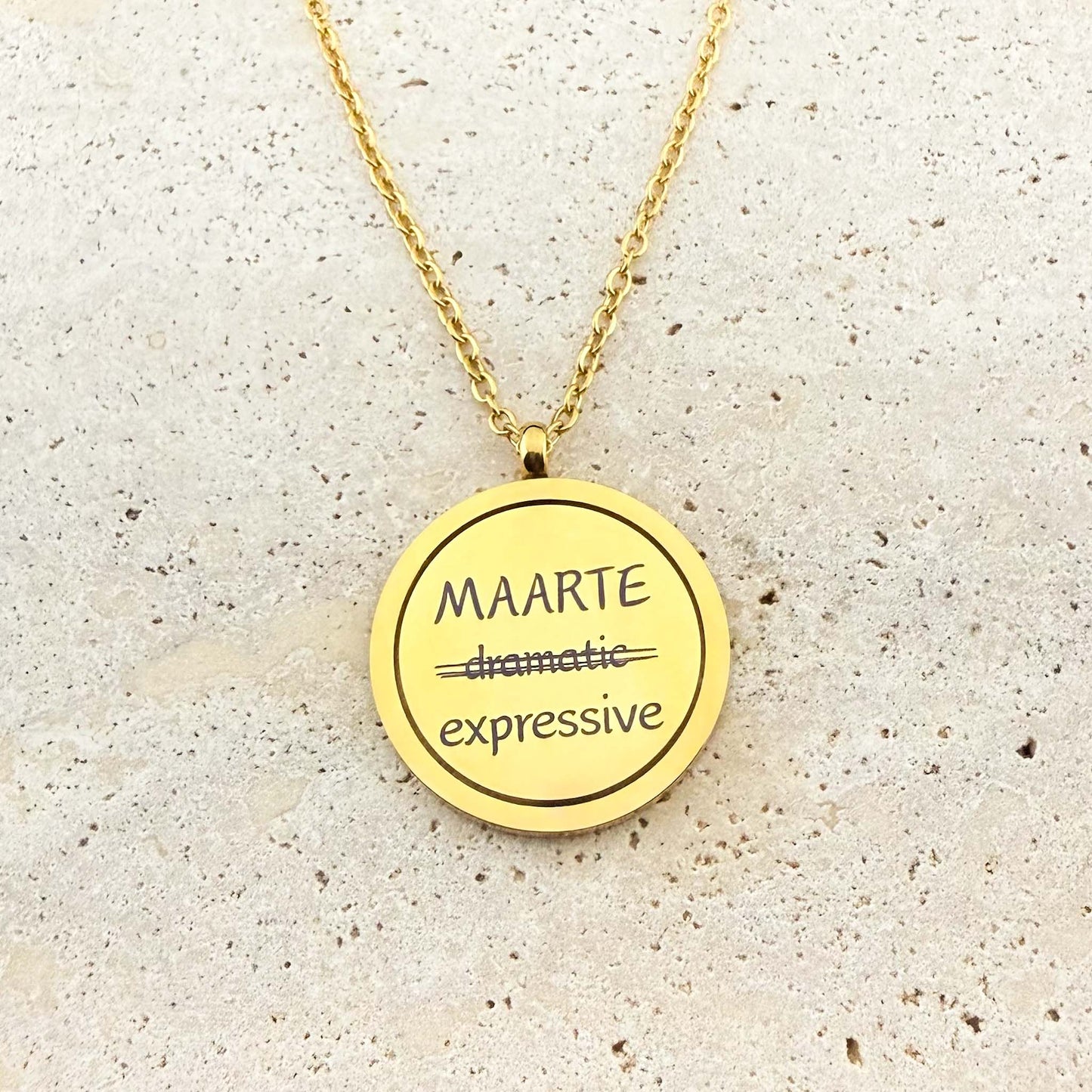 A close-up of an Aking Sinta gold spinning coin fidget necklace featuring the reclaimed Tagalog word "MAARTE." The engraving shows the word "dramatic" crossed out and replaced with the empowering definition "expressive." The circular gold pendant hangs from a cable chain on a light stone surface