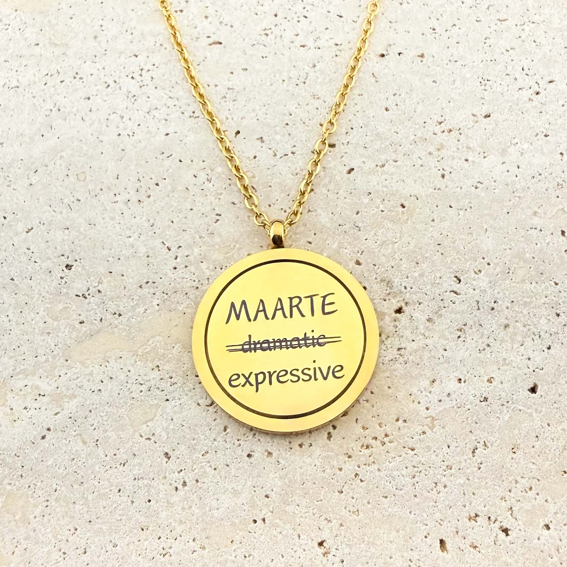 A close-up of an Aking Sinta gold spinning coin fidget necklace featuring the reclaimed Tagalog word "MAARTE." The engraving shows the word "dramatic" crossed out and replaced with the empowering definition "expressive." The circular gold pendant hangs from a cable chain on a light stone surface