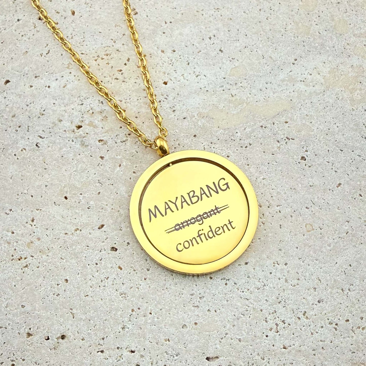 A close-up of an Aking Sinta gold spinning coin fidget necklace featuring the reclaimed Tagalog word "MAYABANG." The engraving shows the word "arrogant" crossed out and replaced with the empowering definition "confident." The circular gold pendant hangs from a polished cable chain and is displayed on a light, textured stone surface