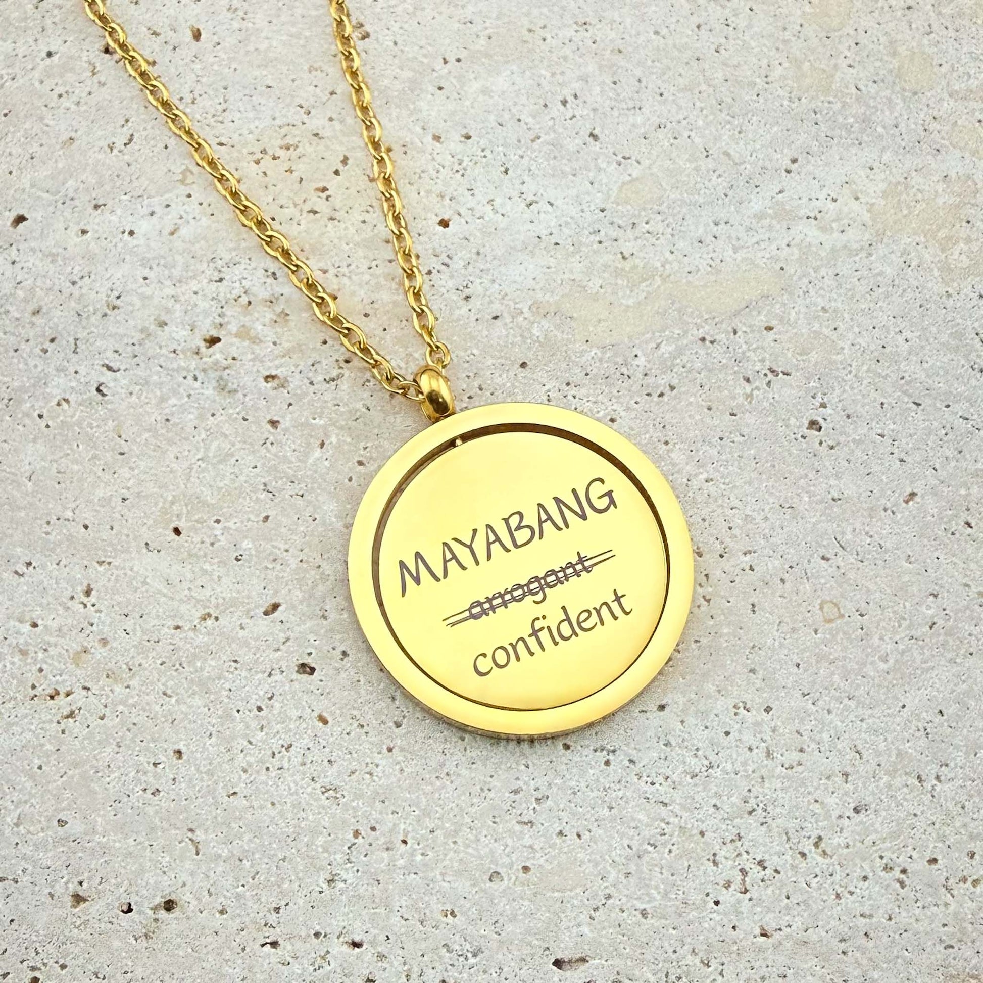 A close-up of an Aking Sinta gold spinning coin fidget necklace featuring the reclaimed Tagalog word "MAYABANG." The engraving shows the word "arrogant" crossed out and replaced with the empowering definition "confident." The circular gold pendant hangs from a polished cable chain and is displayed on a light, textured stone surface