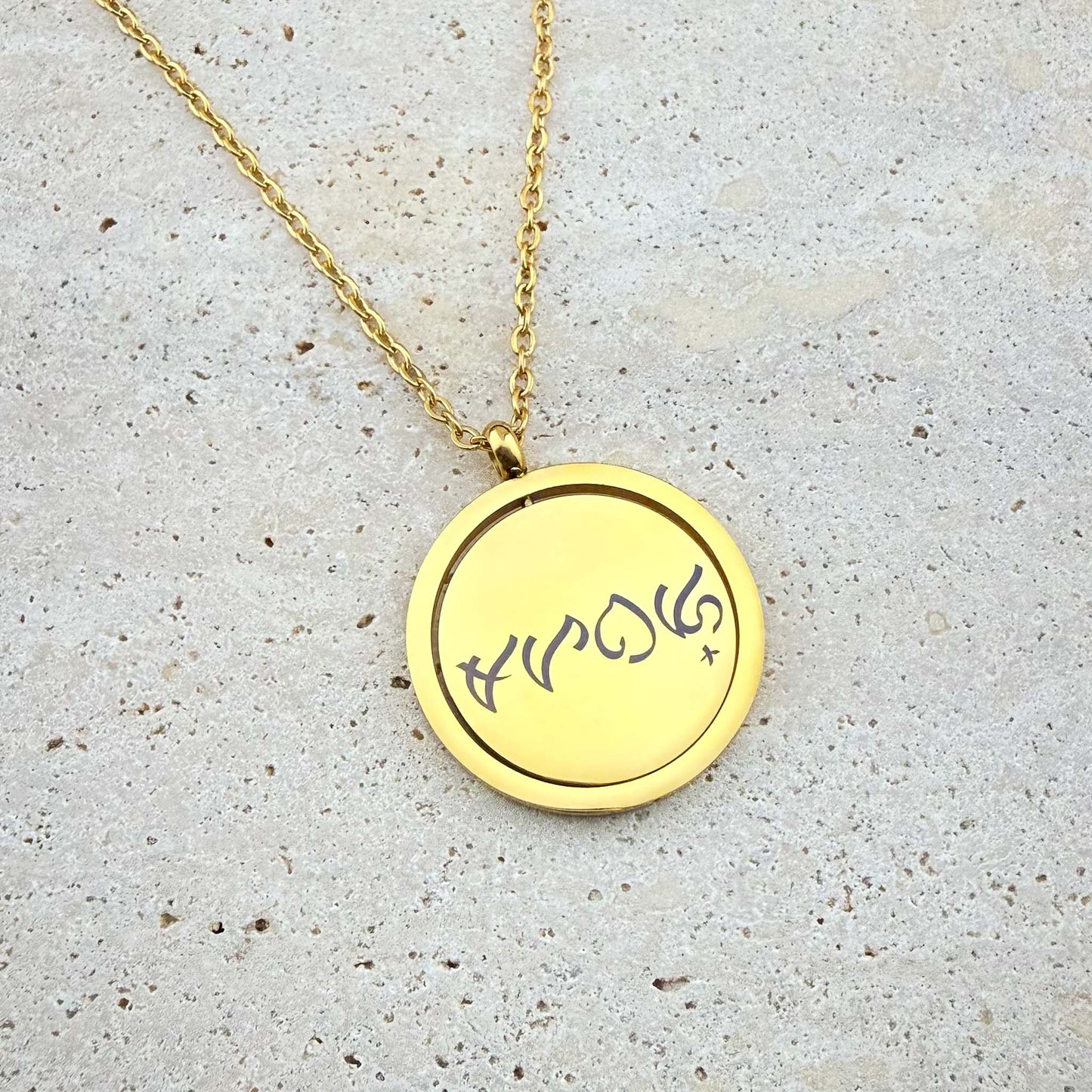 A close-up of an Aking Sinta gold spinning coin fidget necklace featuring a reclaimed Tagalog word engraved in ancient Filipino Baybayin script. The circular gold pendant, designed for tactile relief, hangs from a polished gold cable chain and is displayed on a light, textured stone surface.