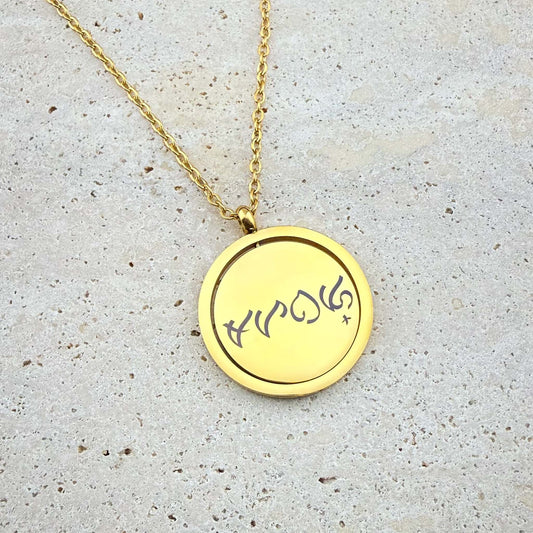 A close-up of an Aking Sinta gold spinning coin fidget necklace featuring a reclaimed Tagalog word engraved in ancient Filipino Baybayin script. The circular gold pendant, designed for tactile relief, hangs from a polished gold cable chain and is displayed on a light, textured stone surface.