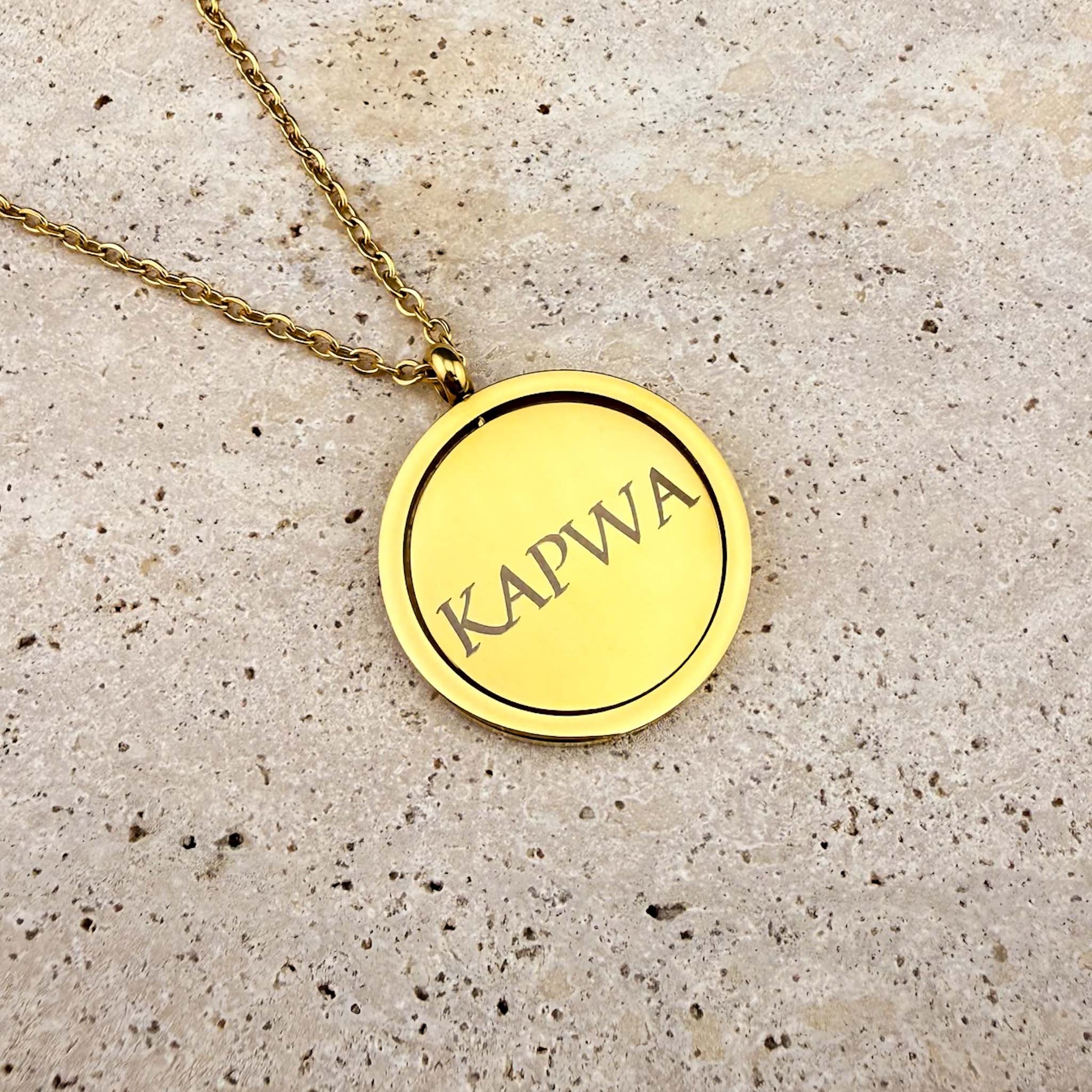 Reverse side of the Aking Sinta gold Spinning Coin necklace engraved with the word "Kapwa" in ancient Baybayin script, displayed on a marble background.