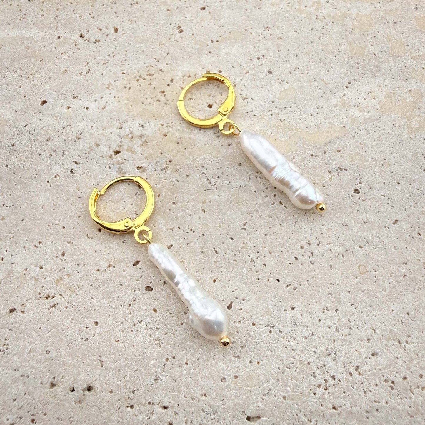 Pair of gold Aking Sinta huggie earrings with long, vertical white stick baroque pearls on a white marble background