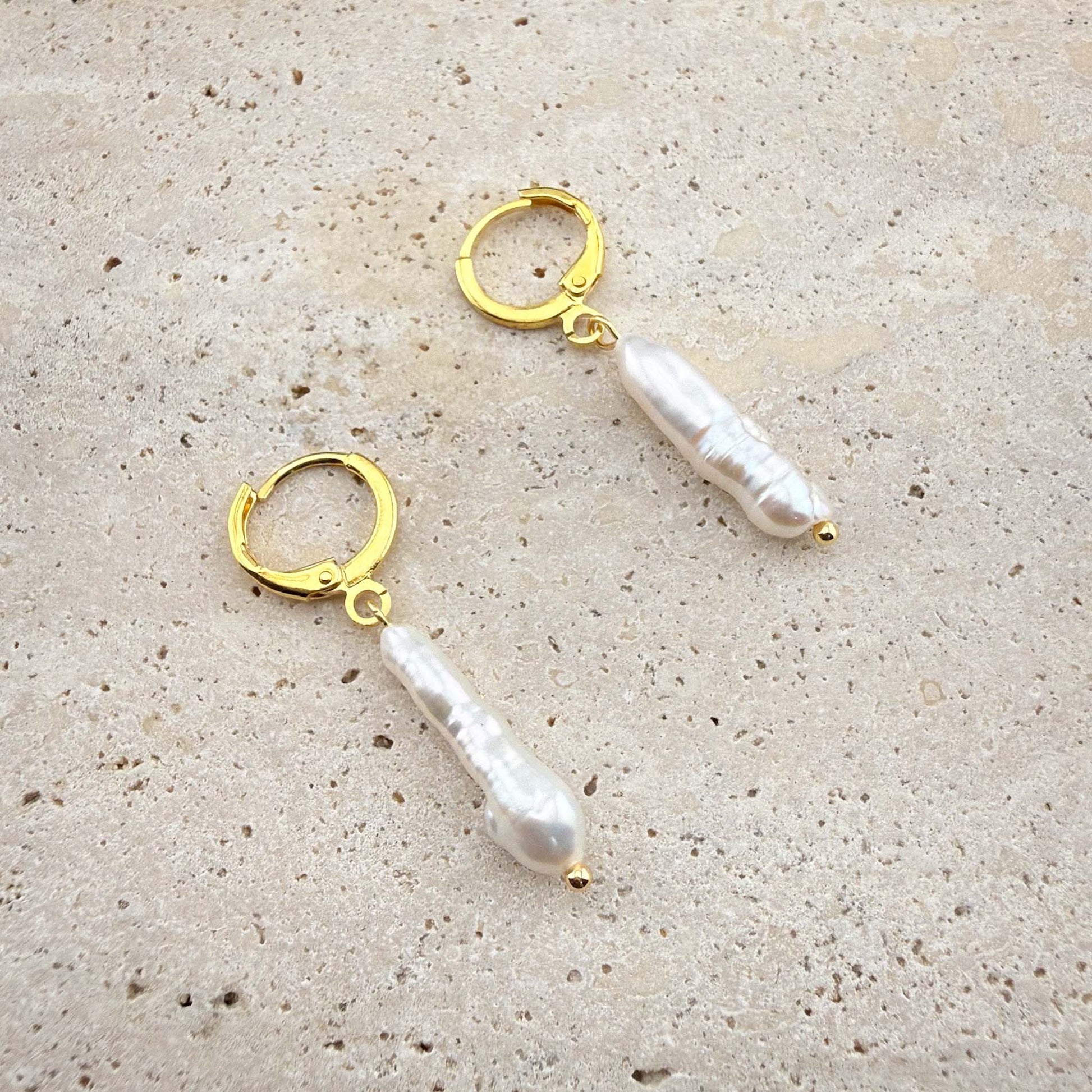Pair of gold Aking Sinta huggie earrings with long, vertical white stick baroque pearls on a white marble background