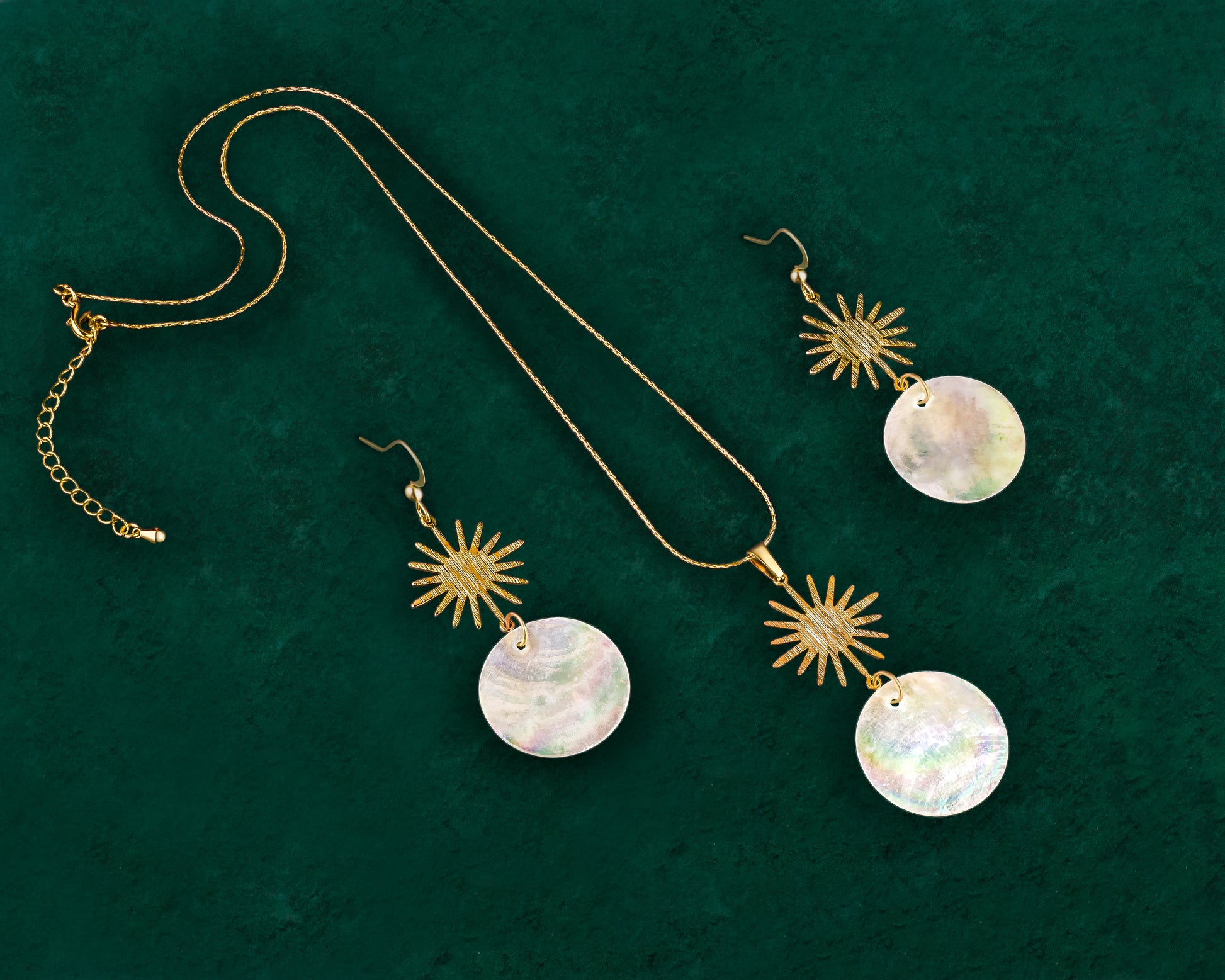 A matching Aking Sinta jewelry set featuring a gold sun motif and iridescent white Capiz shell pendant necklace and a pair of matching drop earrings. Each piece showcases the shimmering natural shell disc suspended from a brushed gold sun design, presented against a dark green velvet background