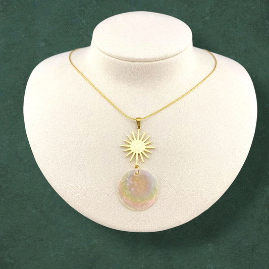 A gold Aking Sinta necklace displayed on a white jewelry mannequin. The pendant features a brushed gold sun motif sitting above a circular, iridescent Capiz shell disc. The piece hangs from a delicate gold chain against a dark green textured background