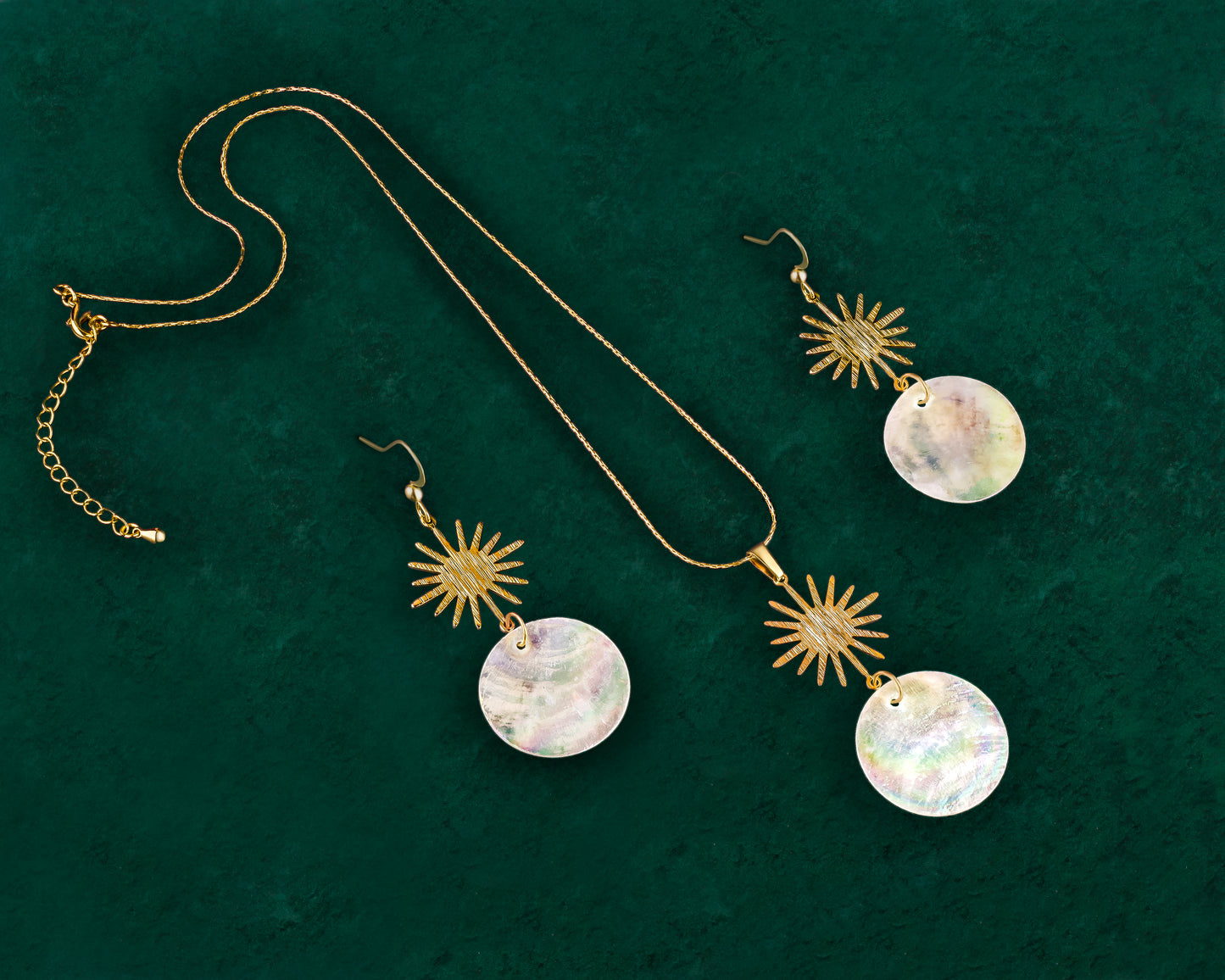 A matching Aking Sinta jewelry set featuring a gold sun motif and iridescent white Capiz shell pendant necklace and a pair of matching drop earrings. Each piece highlights the unique, shimmering, natural shell discs suspended from a brushed gold sun design, presented against a deep green velvet background