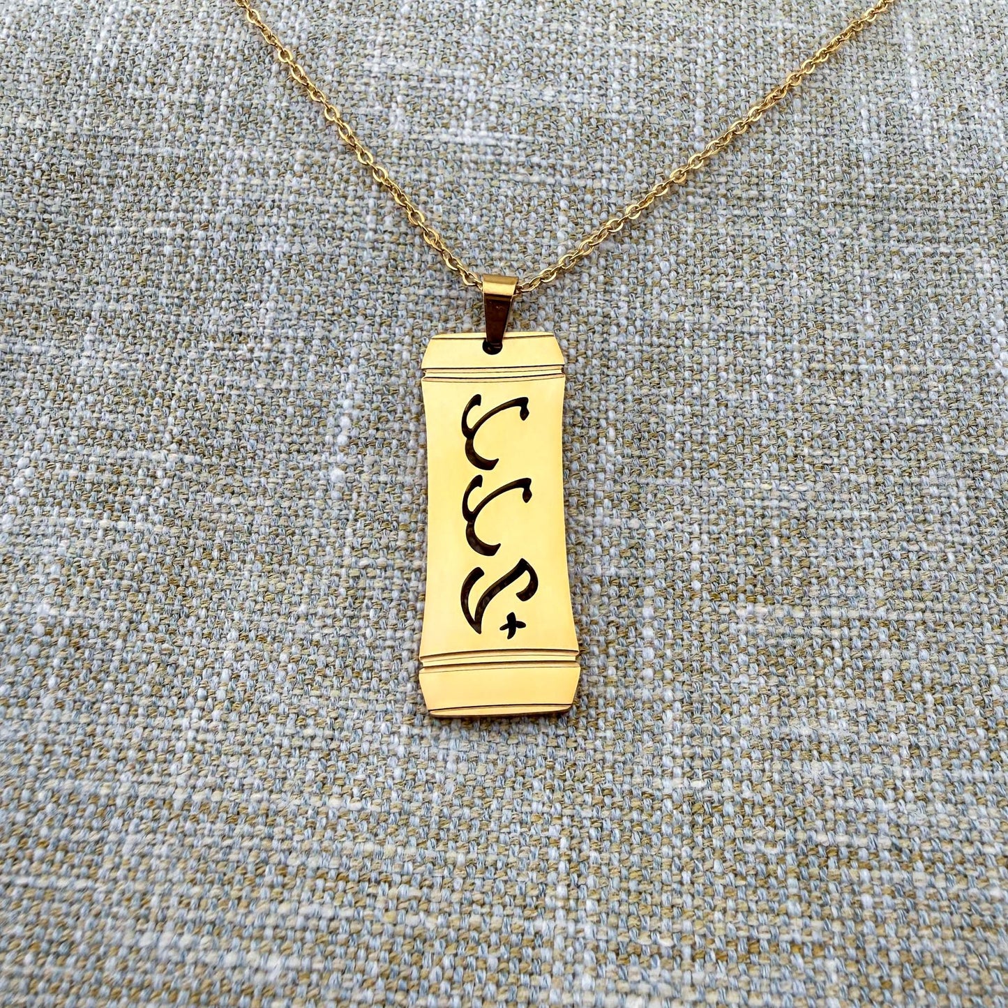 Alt Text:
An Aking Sinta gold vertical pendant necklace centered on a textured blue-gray and gold woven linen background. The matte gold rectangular pendant features a stylized bamboo frame outline with the word "Tatay" (Father) intricately cut out in traditional script-like Baybayin characters, suspended from a fine gold link chain
