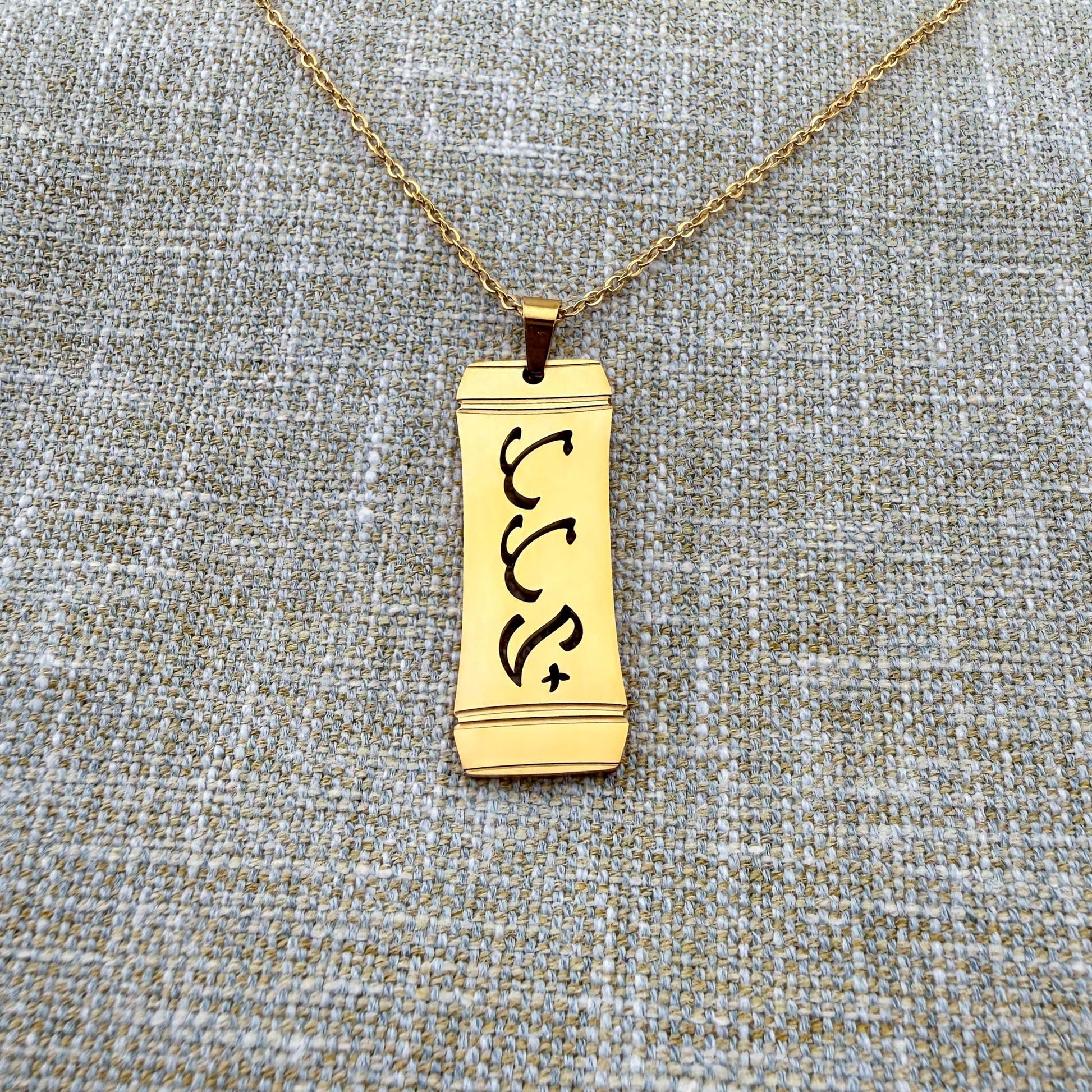 Alt Text:
An Aking Sinta gold vertical pendant necklace centered on a textured blue-gray and gold woven linen background. The matte gold rectangular pendant features a stylized bamboo frame outline with the word "Tatay" (Father) intricately cut out in traditional script-like Baybayin characters, suspended from a fine gold link chain