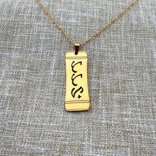 Alt Text:
An Aking Sinta gold vertical pendant necklace centered on a textured blue-gray and gold woven linen background. The matte gold rectangular pendant features a stylized bamboo frame outline with the word "Tatay" (Father) intricately cut out in traditional script-like Baybayin characters, suspended from a fine gold link chain