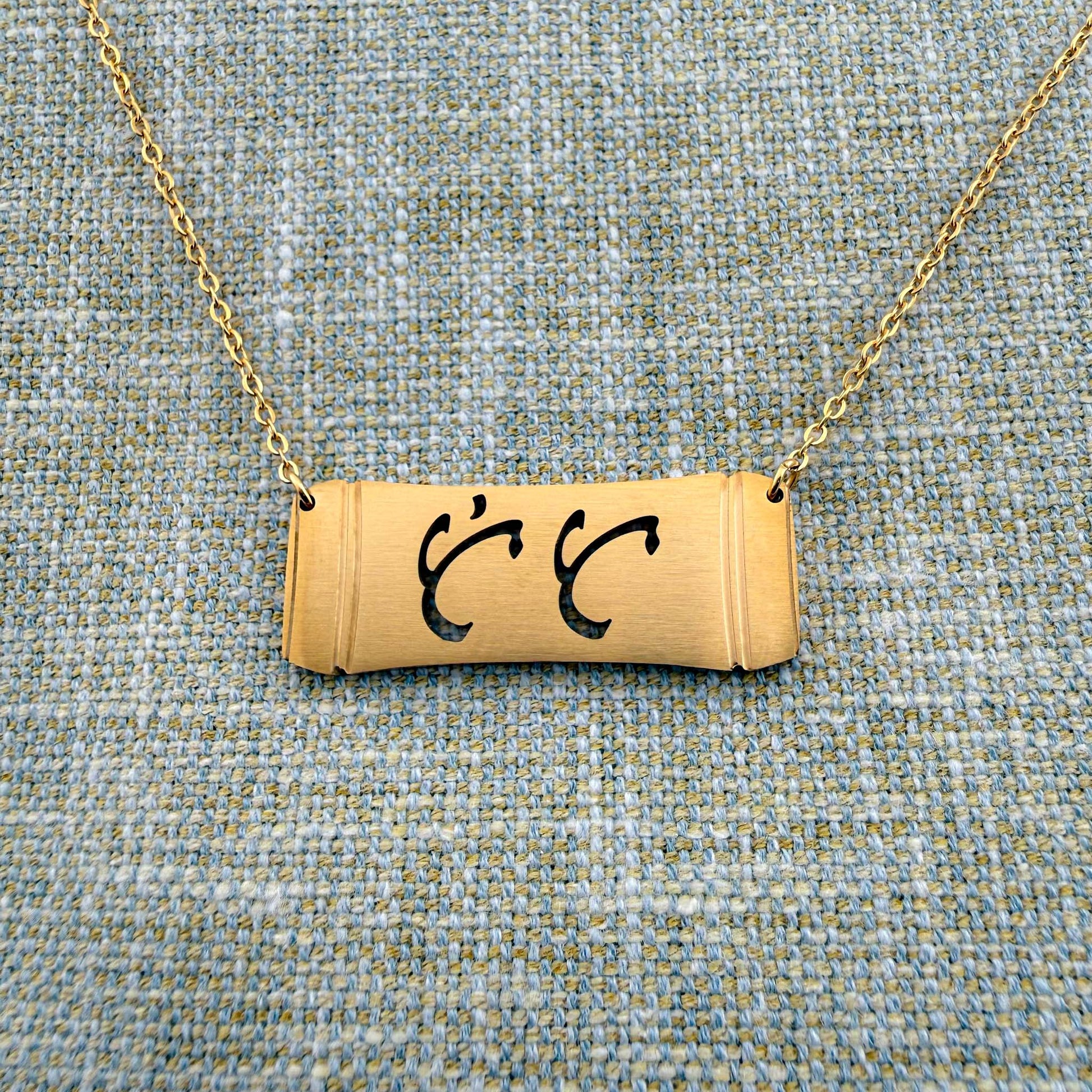 An Aking Sinta gold horizontal bar necklace centered on a textured blue and gold woven linen background. The matte gold pendant features a stylized bamboo section silhouette with the word "Tita" (Aunt) cleanly cut out in elegant, traditional Baybayin script characters, suspended from a fine gold link chain.
