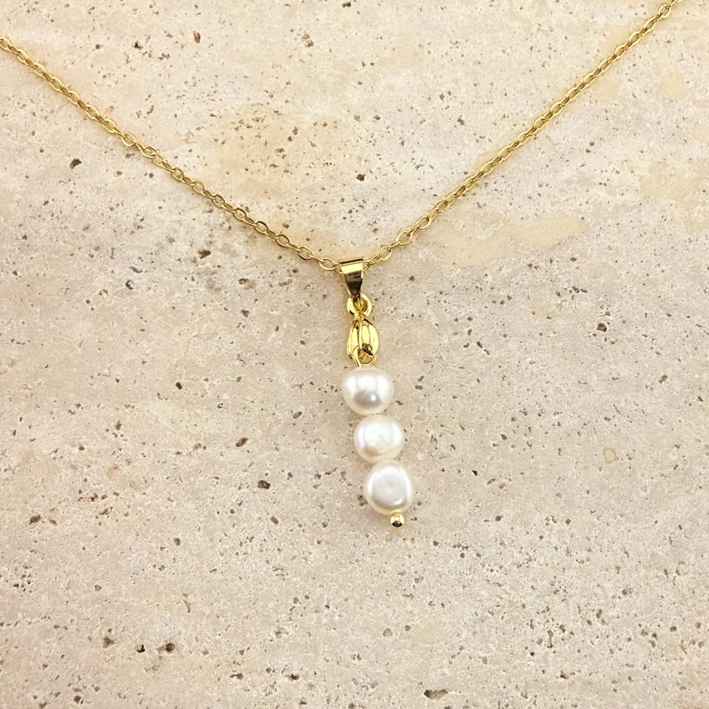 A gold Aking Sinta necklace featuring a delicate pendant composed of three white, organic-shaped baroque pearls stacked vertically. The pearls hang from a polished gold bail and a fine gold cable chain, presented on a light, textured natural stone surface