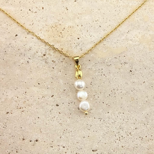A gold Aking Sinta necklace featuring a delicate pendant composed of three white, organic-shaped baroque pearls stacked vertically. The pearls hang from a polished gold bail and a fine gold cable chain, presented on a light, textured natural stone surface