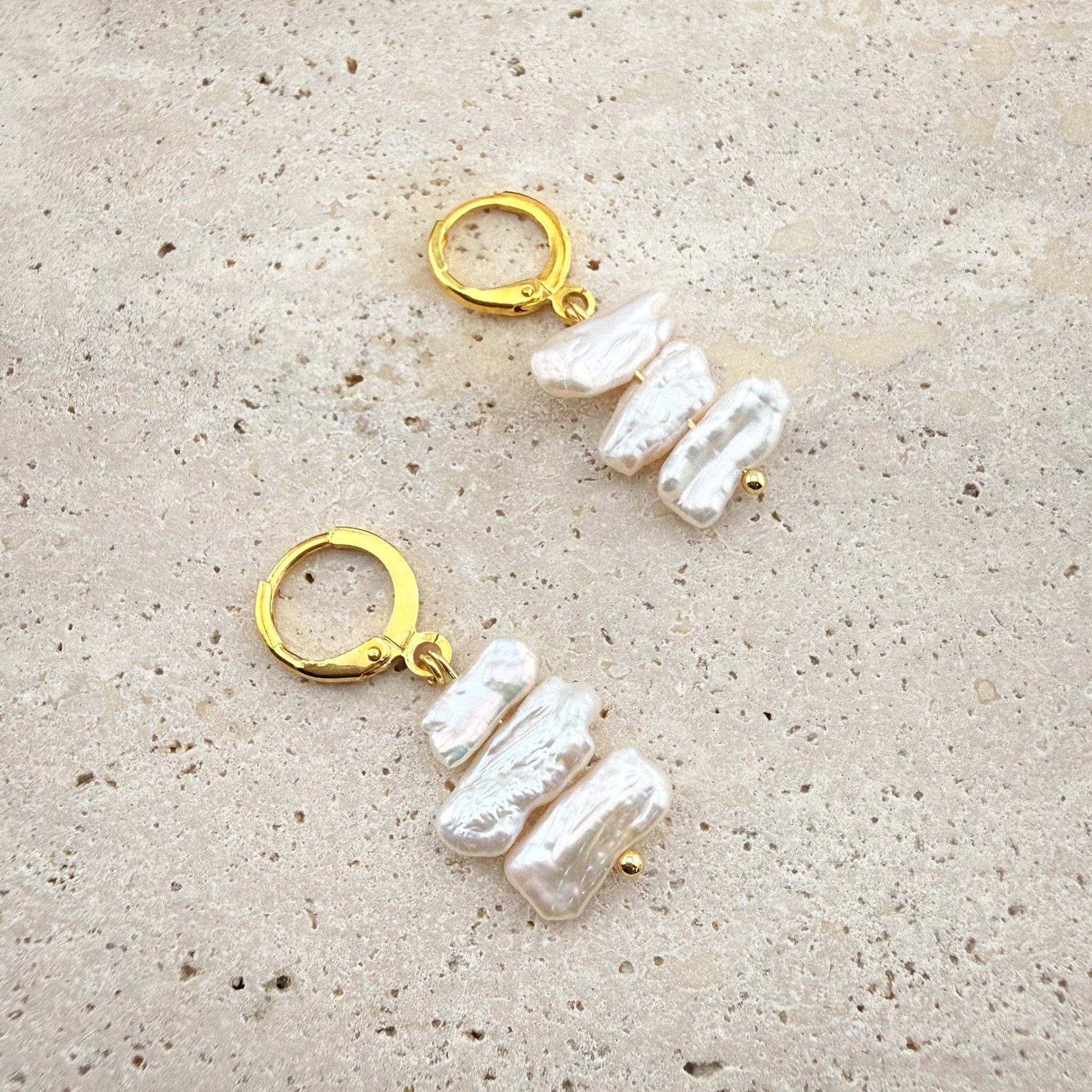 Pair of Aking Sinta gold huggie earrings with three cascading, flat white Keshi pearls on a white marble surface.