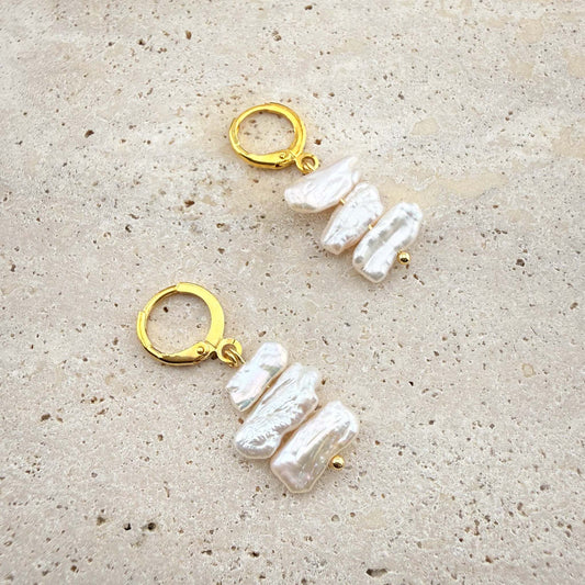 Pair of Aking Sinta gold huggie earrings with three cascading, flat white Keshi pearls on a white marble surface.