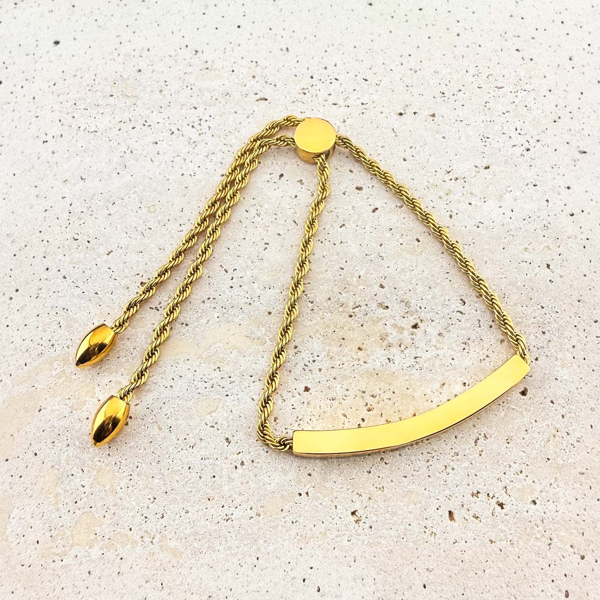 A gold Aking Sinta twisted rope chain bracelet with a polished, curved bar ready for bespoke engraving. The adjustable bolo-style bracelet features a circular slider clasp and elegant teardrop end charms, displayed on a light-colored natural stone surface