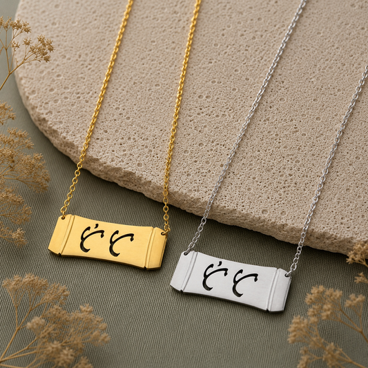 A product lifestyle photograph featuring two Aking Sinta horizontal bamboo bar necklaces in matte gold and silver finishes side by side. The parallel curved bar pendants display the word "Tita" (Aunt) in elegant cutout Baybayin script characters, arranged over a light beige textured stone slab and sage green fabric backdrop with dried baby's breath