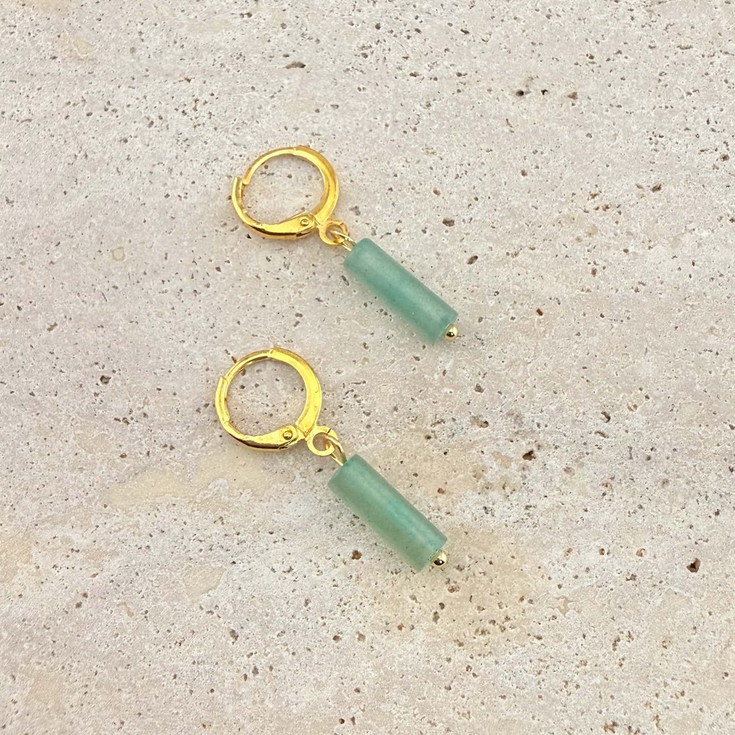 A pair of Aking Sinta gold huggie hoop earrings featuring cylinder-shaped green aventurine column pendants. These natural gemstone drop earrings are displayed on a light sand-colored stone surface