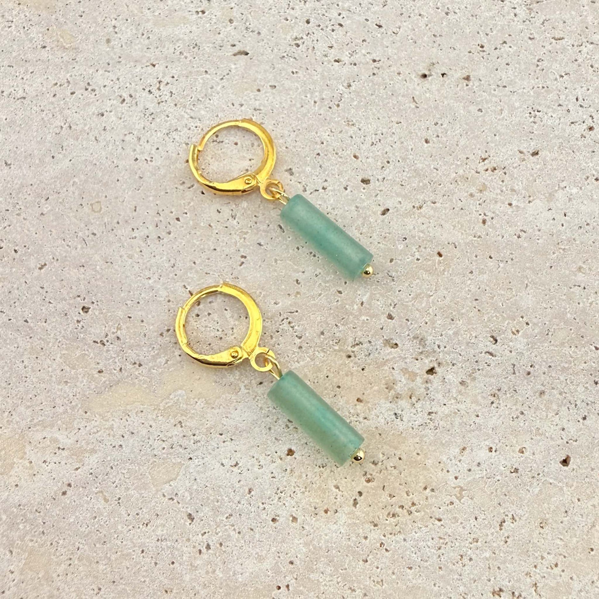 A pair of Aking Sinta gold huggie hoop earrings featuring cylinder-shaped green aventurine column pendants. These natural gemstone drop earrings are displayed on a light sand-colored stone surface