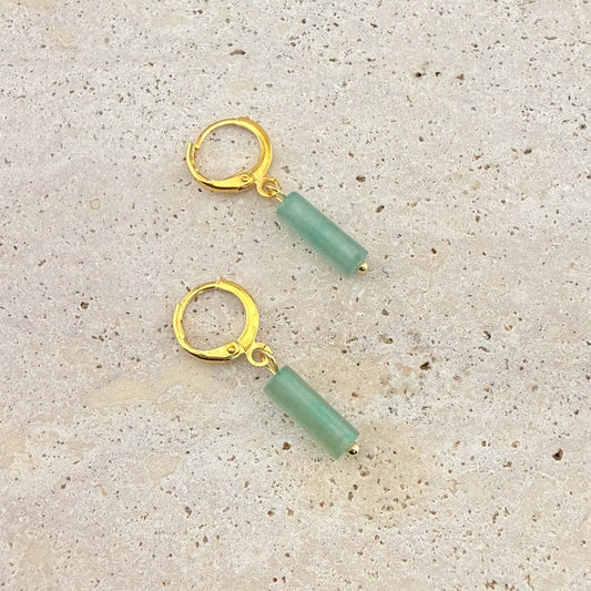 A pair of Aking Sinta gold huggie hoop earrings featuring cylinder-shaped green aventurine column pendants. These natural gemstone drop earrings are displayed on a light sand-colored stone surface
