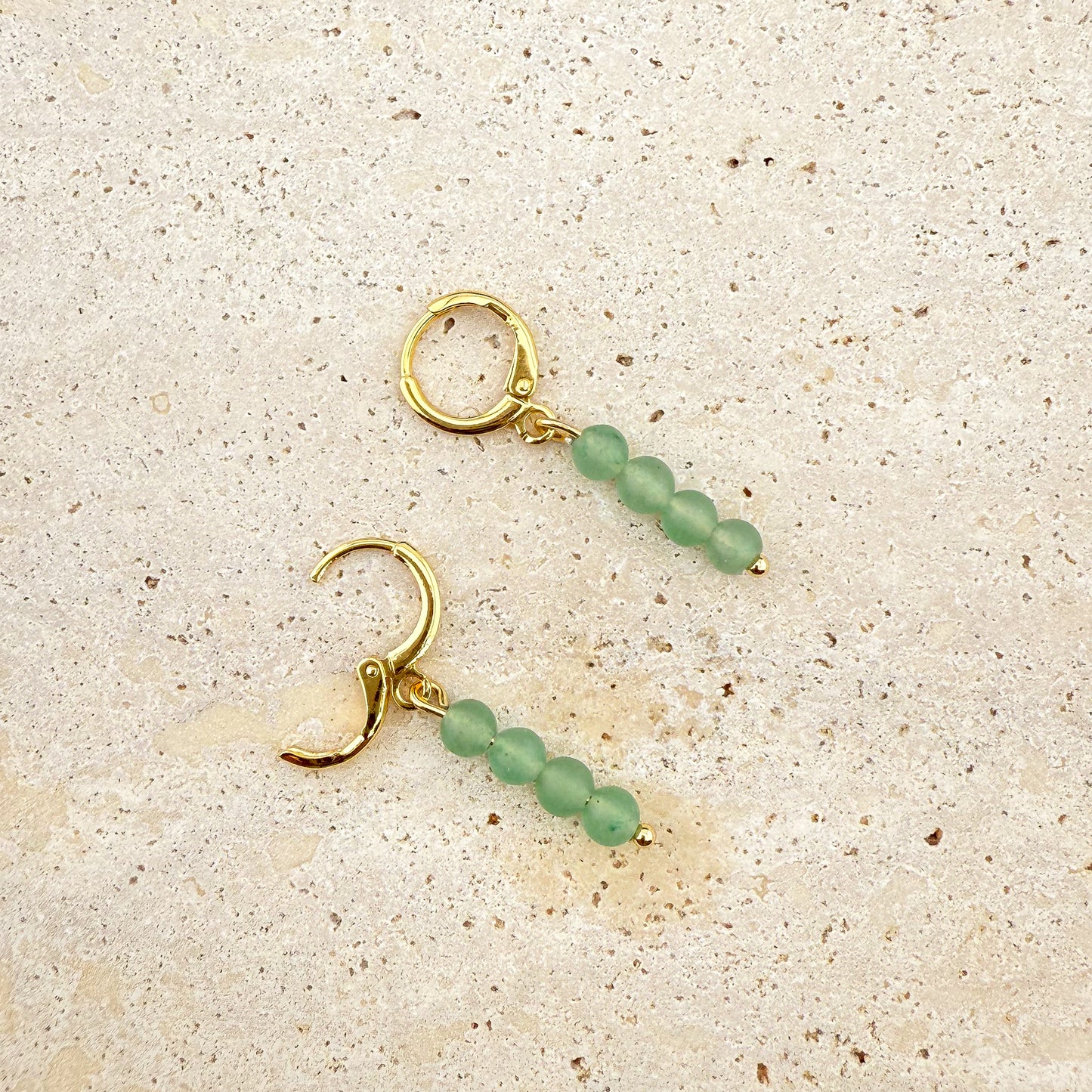 A close-up of the Aking Sinta Green Aventurine Huggies on a white marble surface; one earring is closed, while the other has its polished gold clasp open, showing the hinge and the small, faceted green gemstone drop.