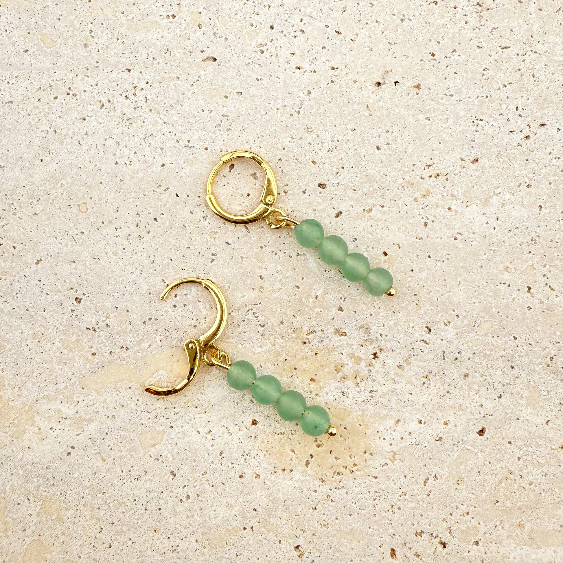 A close-up of the Aking Sinta Green Aventurine Huggies on a white marble surface; one earring is closed, while the other has its polished gold clasp open, showing the hinge and the small, faceted green gemstone drop.