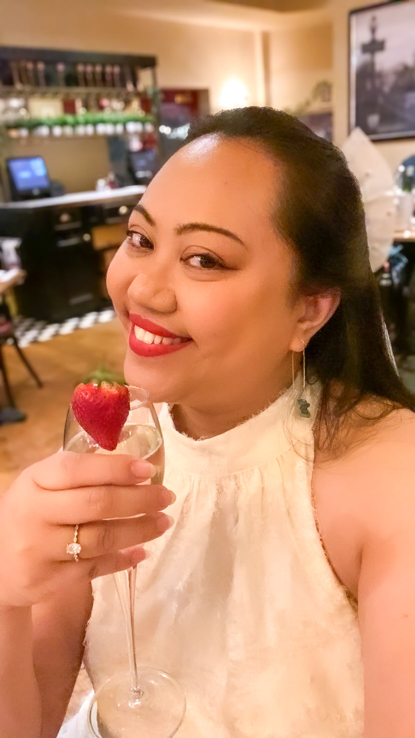 Lifestyle photo of a woman in a white dress wearing Aking Sinta gold drop earrings with clustered green aventurine nuggets; she is holding a champagne glass against a dark green background, highlighting the jewelry's elegant and celebratory appeal.