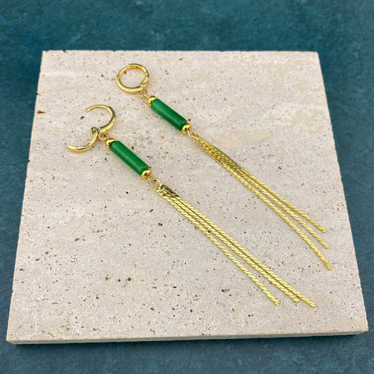 A pair of Aking Sinta gold tassel earrings on a gray marble slab, featuring a vertical column of dark green jade suspended above a fringe of delicate gold chains.