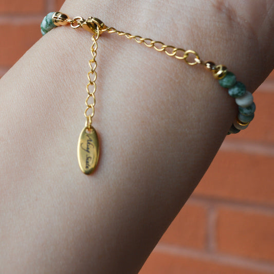 Close-up of the back of a Green Spot Jasper bracelet worn on a wrist, featuring the gold extender chain and a small circular metal tag with the "Aking Sinta" brand name engraved on it