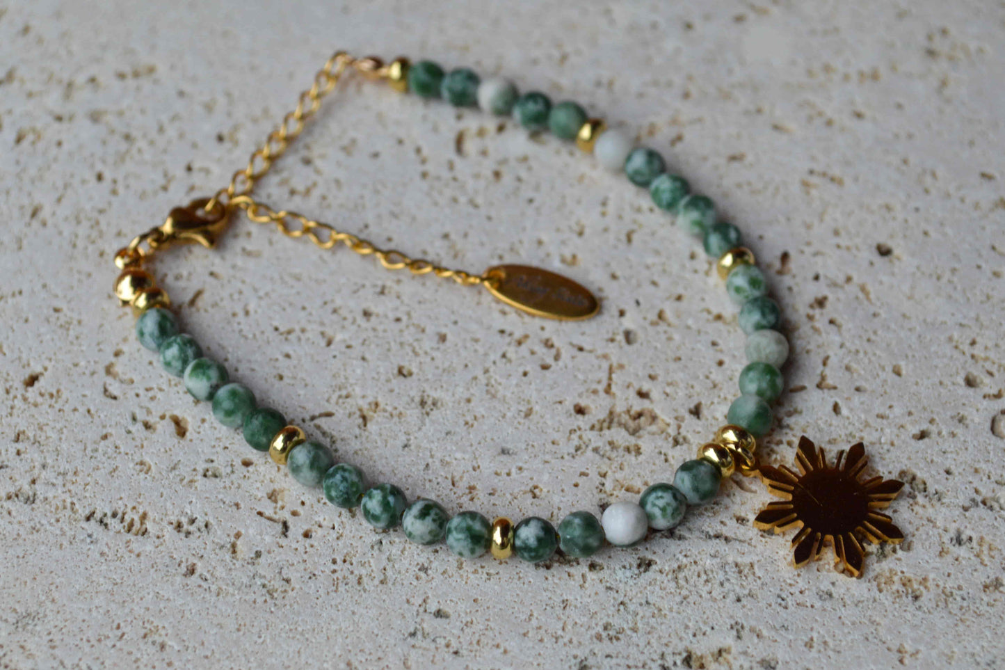 Aking Sinta Green Spot Jasper beaded bracelet laid flat on a white marble surface; featuring earthy green and brown stone beads, a gold adjustable extender chain, and a polished gold circular charm engraved with the brand name.