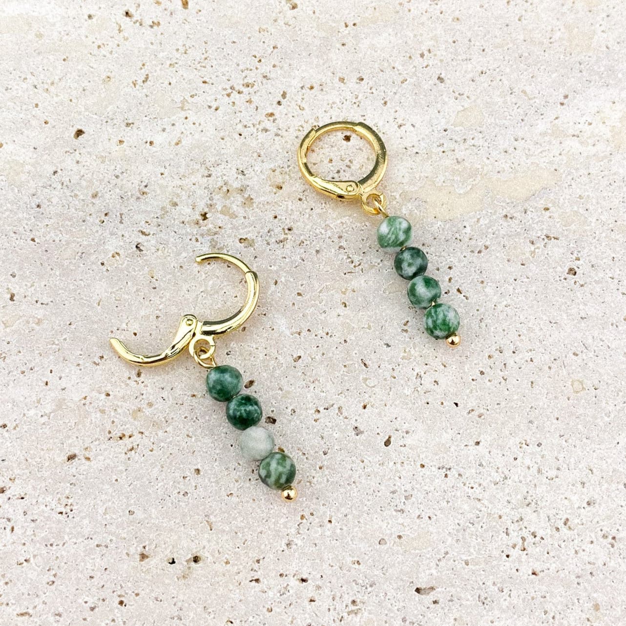 A close-up of Aking Sinta Green Spot Jasper Huggies on a textured gray rock surface; one gold earring is snapped shut while the other is open, showing the hinge mechanism and the dangling speckled green stone.