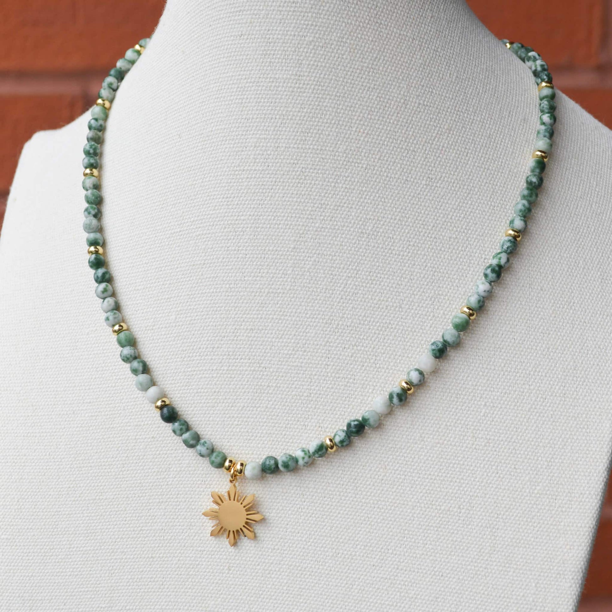 Close-up of a Green Spot Jasper beaded necklace with a gold 8-ray Araw sun charm, presented on a white professional jewelry display neck; highlighting the natural green stone patterns and Aking Sinta craftsmanship