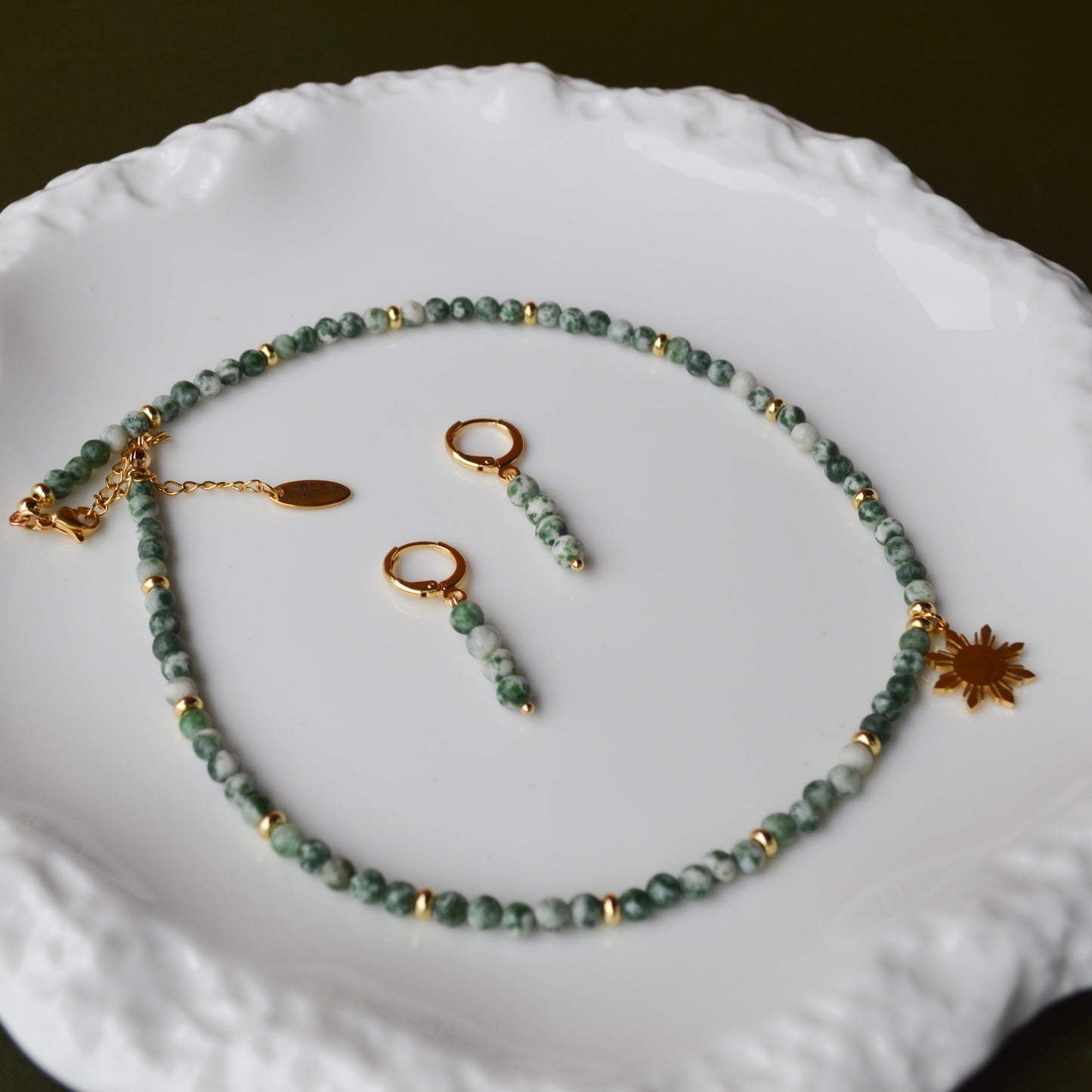 A matching Aking Sinta jewelry set featuring the Green Spot Jasper necklace with an 8-ray Araw charm and gold dainty huggie earrings; displayed on a luxurious iridescent Mother of Pearl surface over a pure white background.