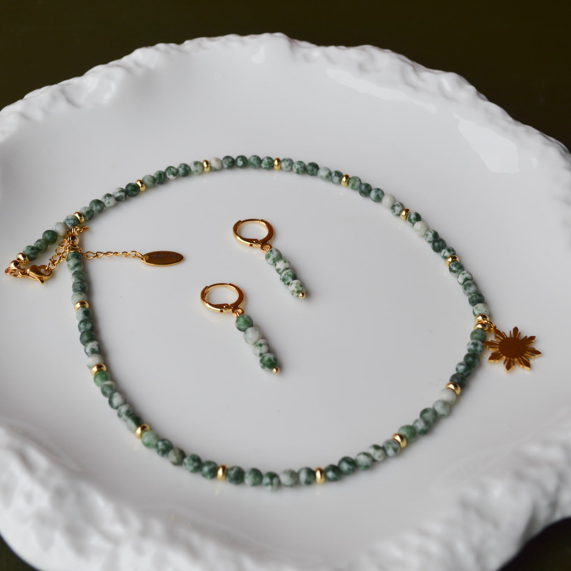 A matching Aking Sinta jewelry set featuring the Green Spot Jasper necklace with an 8-ray Araw charm and gold dainty huggie earrings; displayed on a luxurious iridescent Mother of Pearl surface over a pure white background.