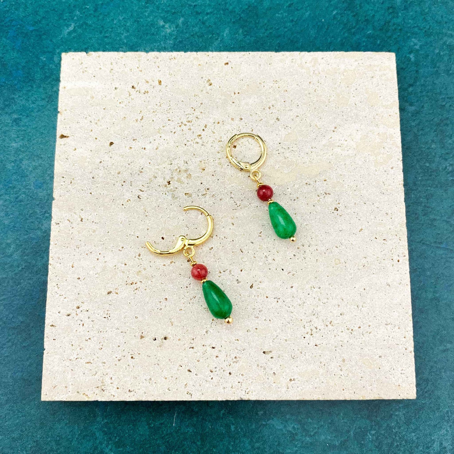 A pair of Aking Sinta gold huggie earrings featuring red jade accents and green jade teardrops, resting on a large, full-size gray marble slab with prominent natural veining.