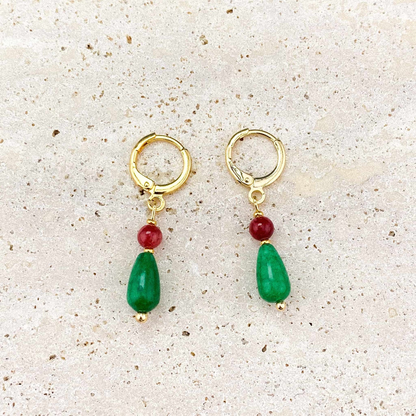 A pair of Aking Sinta gold huggie earrings on a gray marble surface; the design features a small, vibrant Red Jade bead sitting atop a large, smooth Green Jade teardrop charm, inspired by Howl’s iconic jewelry.