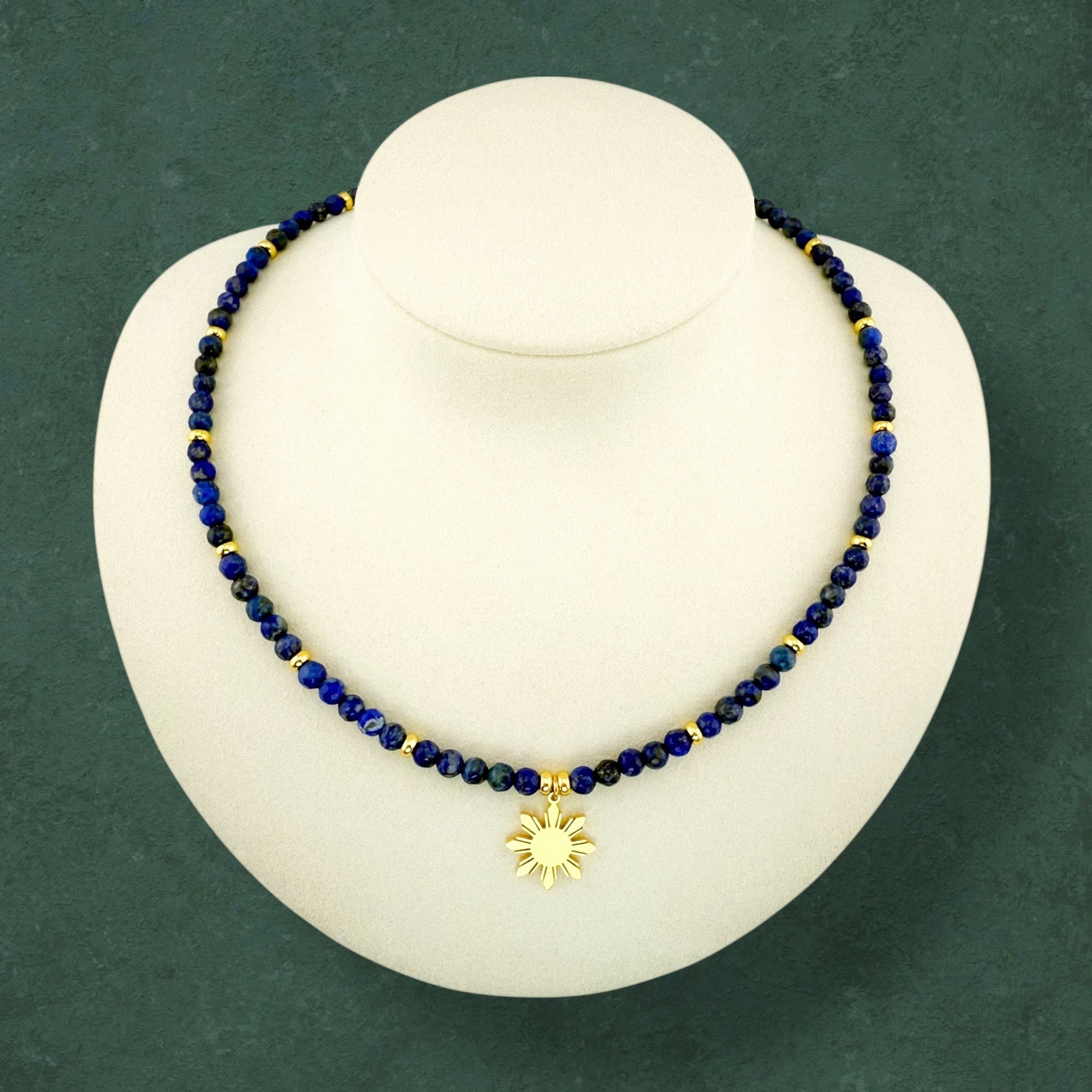 Aking Sinta Lapis Lazuli beaded necklace with a central gold 8-ray Araw sun pendant; the deep blue stones with natural gold pyrite flecks are displayed on a dark green background.