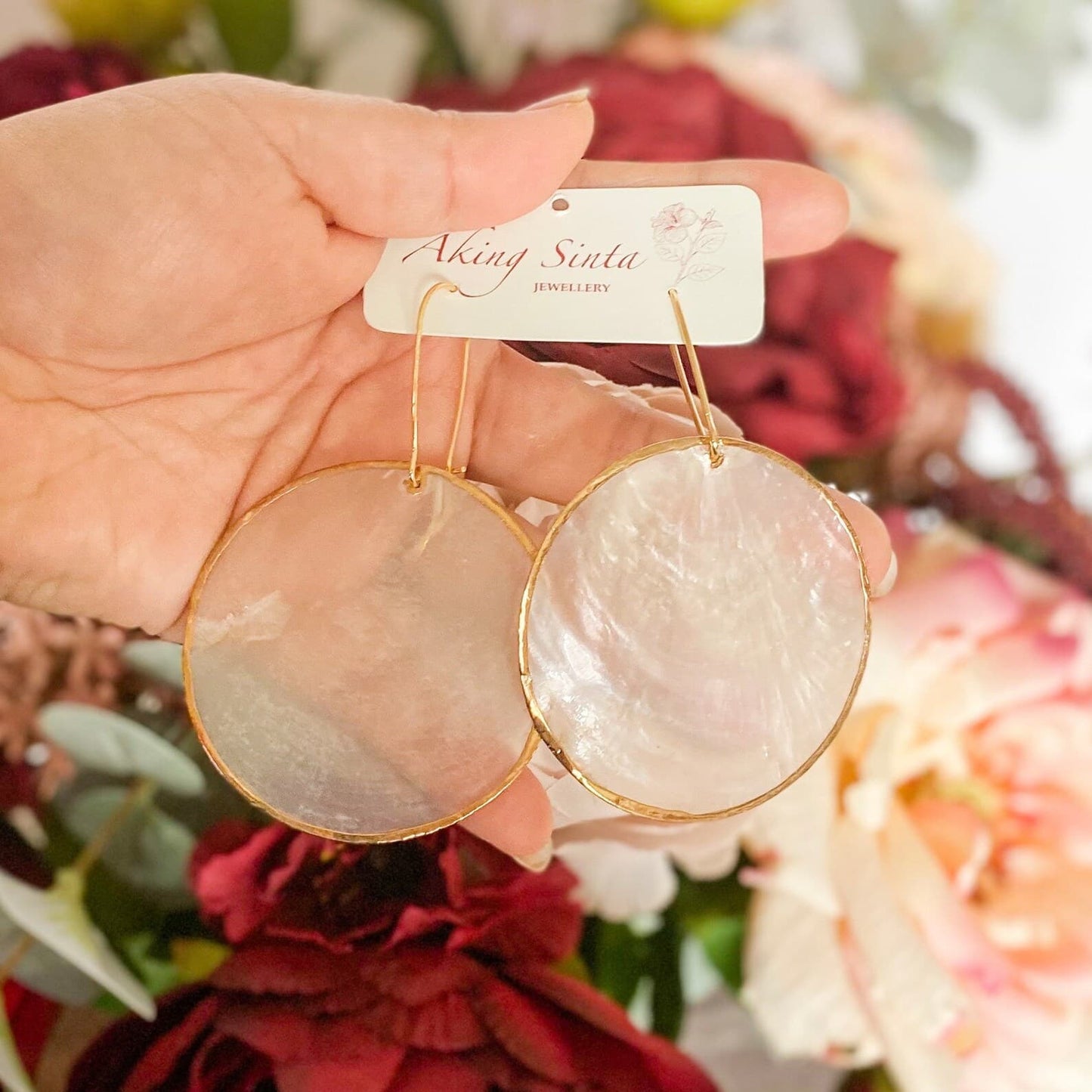 A person’s hand holding up a large, round Aking Sinta Gold Lined Capiz Shell earring; the oversized iridescent white disk is framed by a polished gold metal trim, set against a romantic, soft-focus background of vibrant red roses.