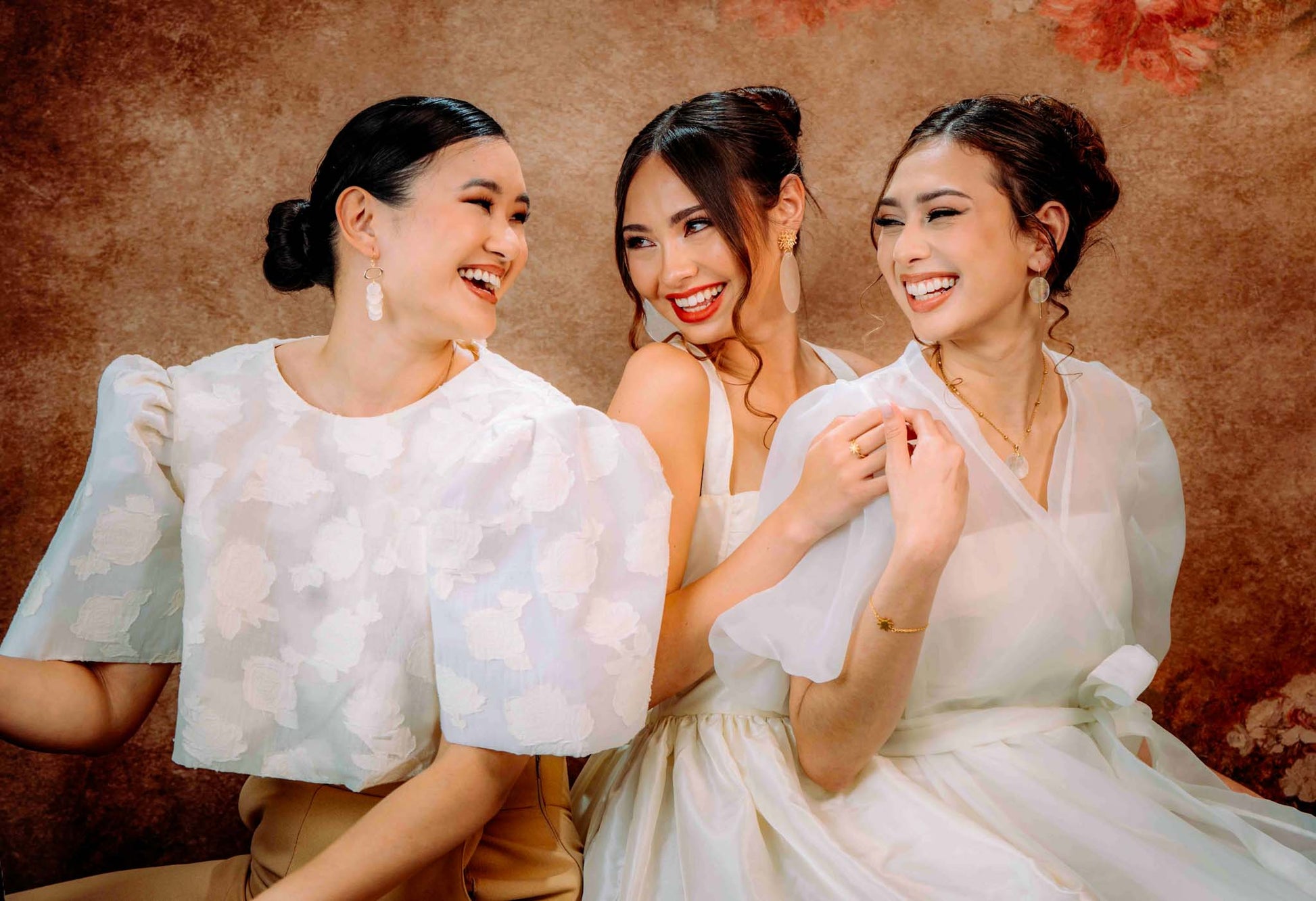 A vibrant lifestyle campaign portrait featuring three smiling women styled in elegant, modern Filipiniana-inspired attire. The models are wearing various pieces from the Aking Sinta collection, including delicate drop earrings and necklaces featuring iridescent Capiz shell accents. The studio setting has a warm, vintage-inspired textured background, emphasizing the timeless, artisanal quality of the jewelry