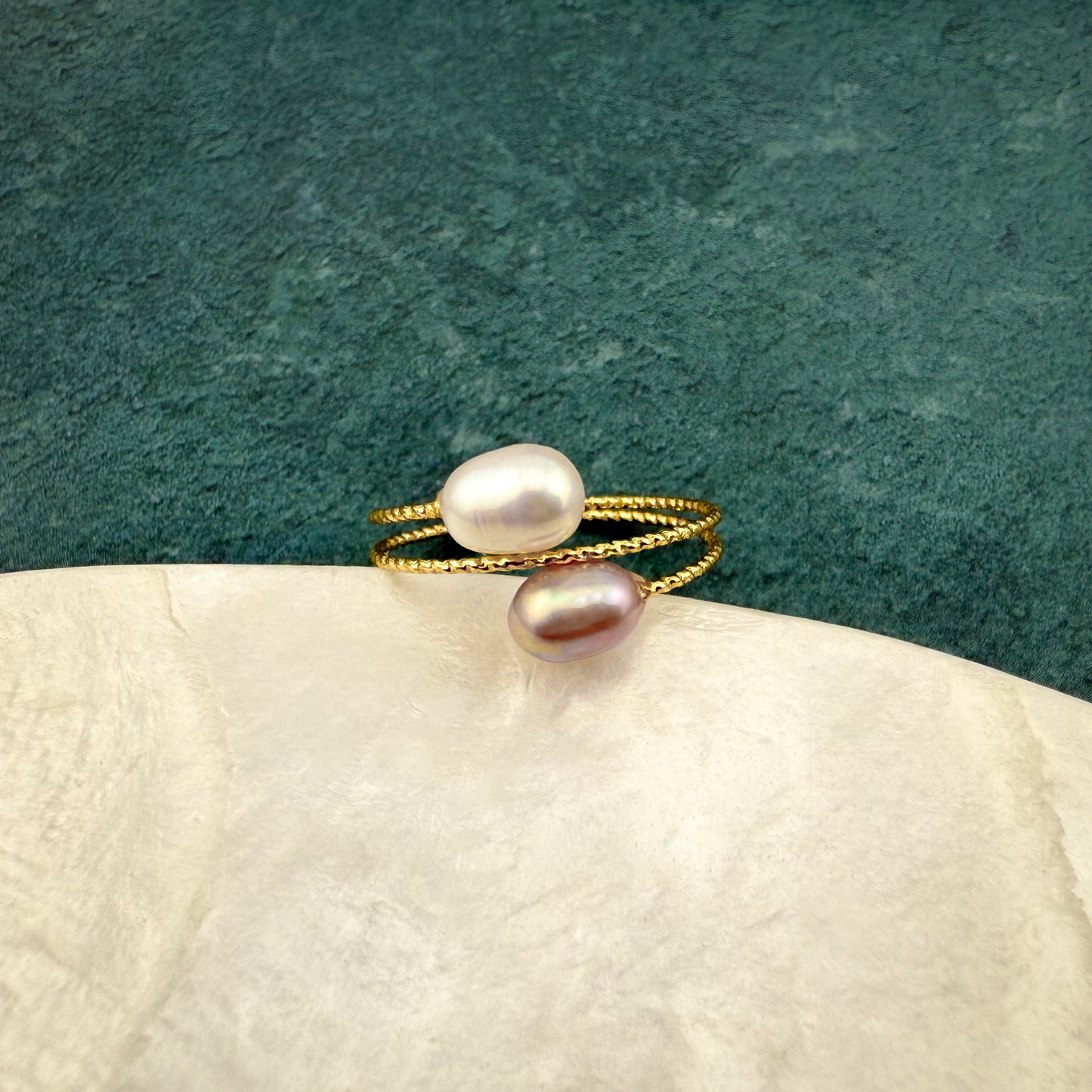 Birds-eye view of an Aking Sinta gold adjustable ring with two pearls, perched on the edge of a white marble disc against a dark green backdrop