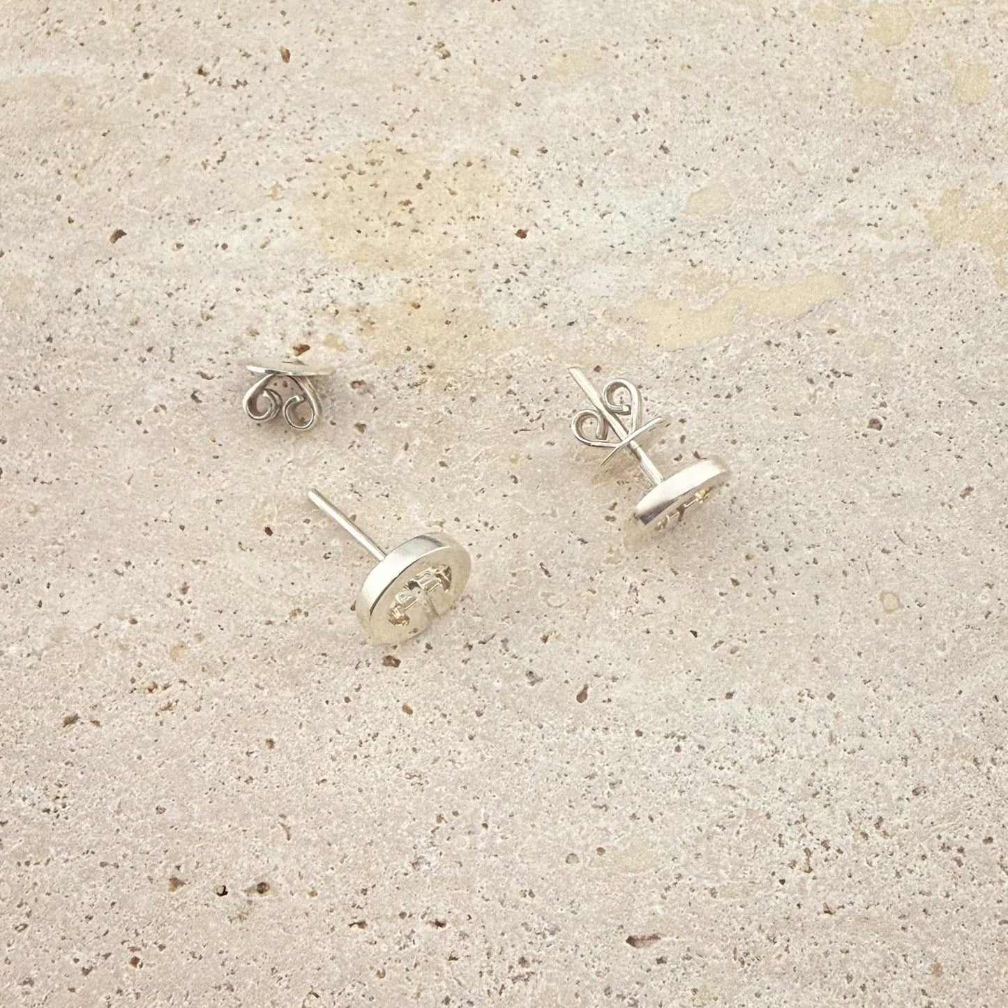A close-up view of Aking Sinta sterling silver Lingling-O stud earrings. The image shows the butterfly scroll backs and post detail of the circular Filipino-inspired studs, displayed on a light natural stone surface