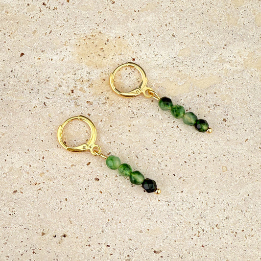 A pair of Aking Sinta gold huggie earrings on a white textured rock; each earring features a vertical drop of four round Malaysian Jade beads in a bright, translucent emerald-green.
